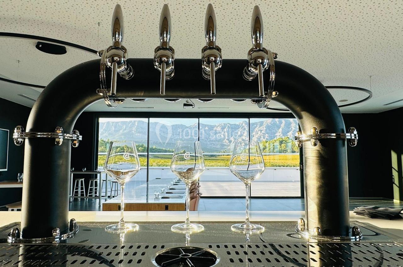 Quatre robinets de distribution de boissons au-dessus de trois verres à pied, avec une vue sur des montagnes et des vignes.