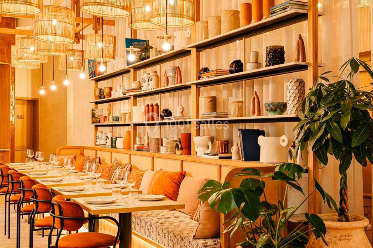 Salle de restaurant lumineuse avec tables alignées, chaises orange, étagères décorées et plantes vertes.