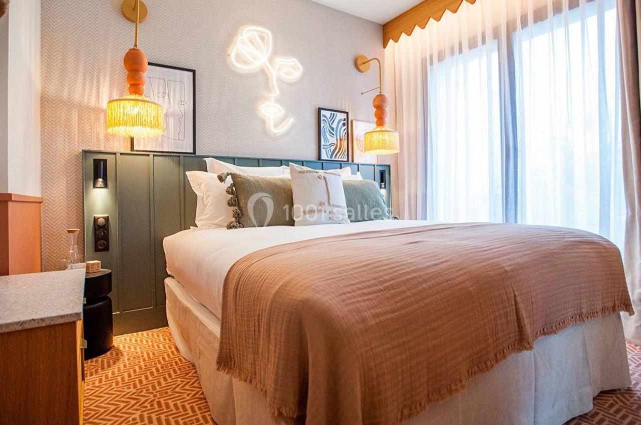 Chambre lumineuse avec un lit double, tête de lit décorée, suspensions en rotin et grande fenêtre avec rideaux translucides.