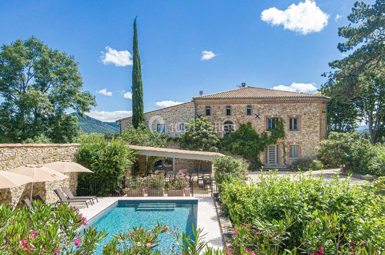 Maison en pierre entourée de végétation avec piscine, terrasse ombragée et ciel bleu en arrière-plan.