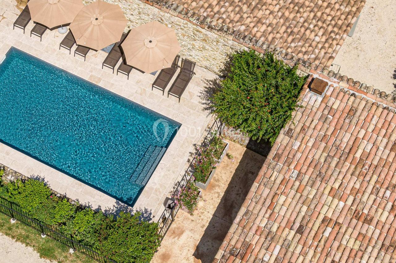 Vue aérienne d'une piscine entourée de chaises longues, parasols, végétation et bâtiments en tuiles.