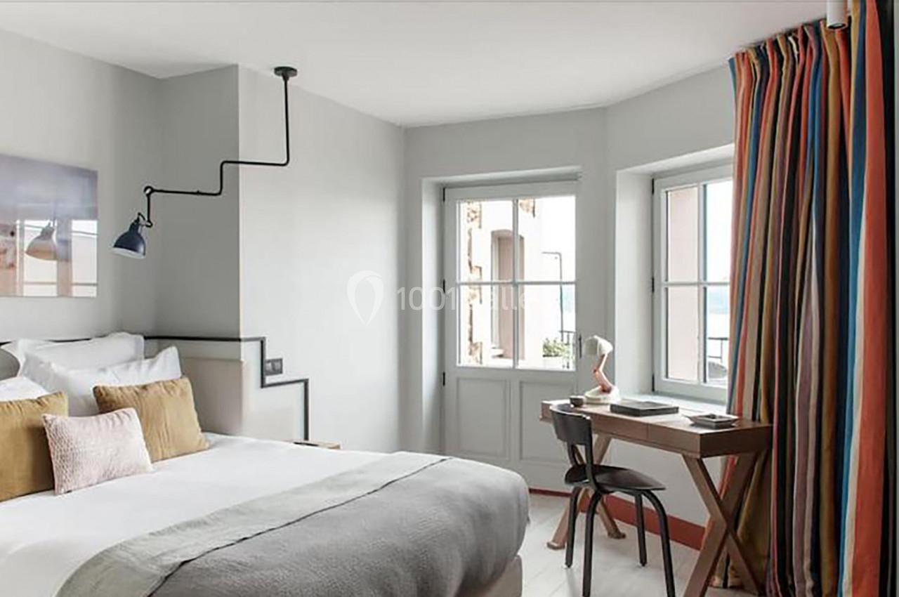 Chambre lumineuse avec lit double, bureau en bois, lampe et rideaux colorés près de fenêtres donnant sur l'extérieur.