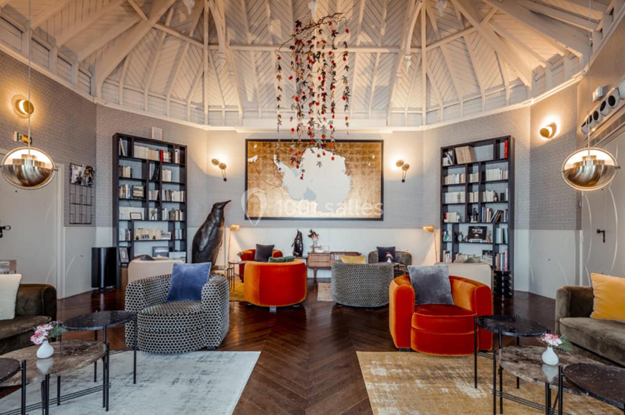 Salon lumineux avec fauteuils colorés, tables basses, étagères remplies de livres et plafond voûté en bois blanc.