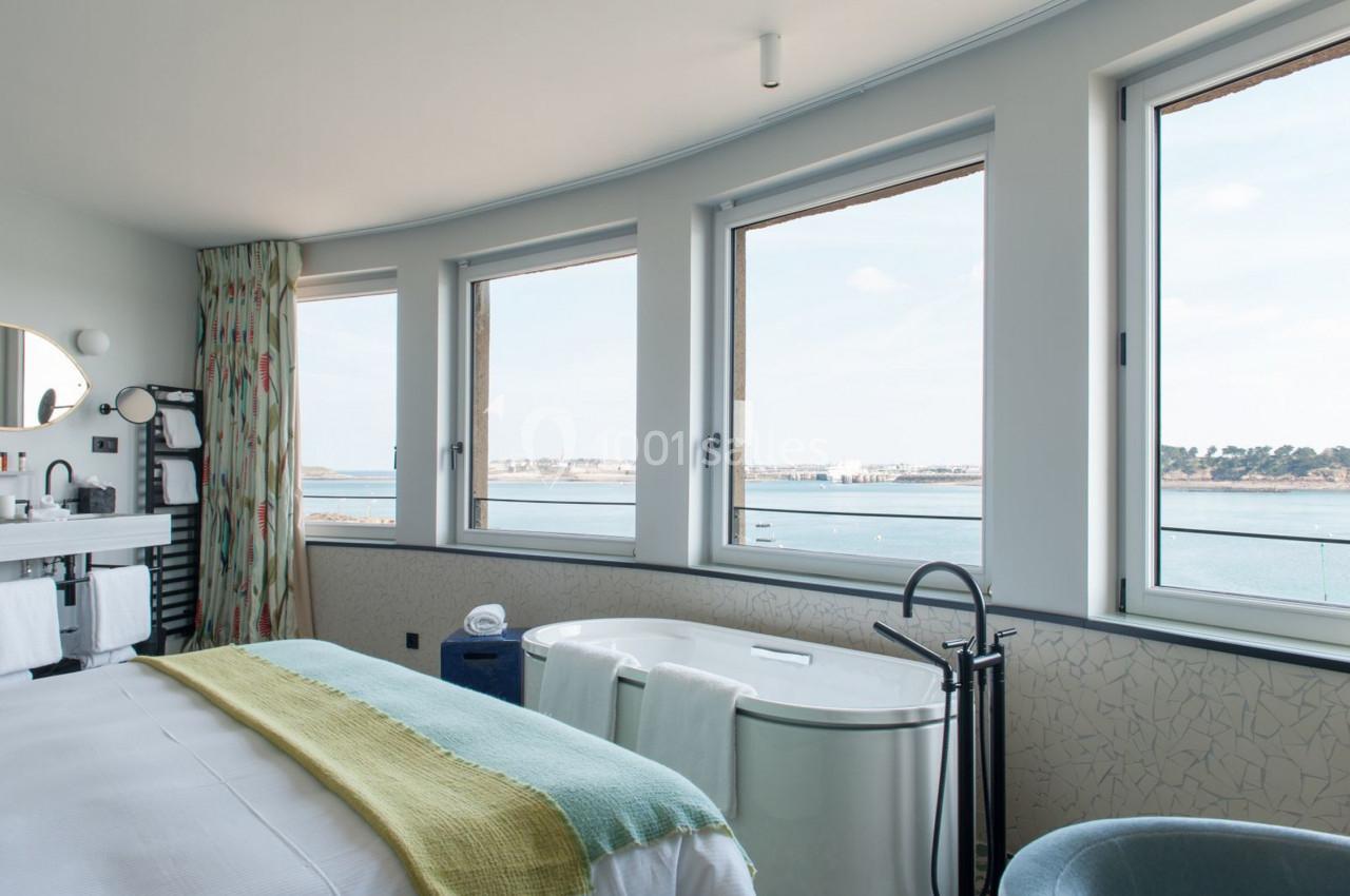 Chambre lumineuse avec lit, baignoire et vue sur la mer à travers de grandes fenêtres.