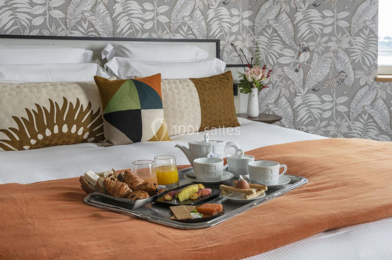 Petit-déjeuner servi sur un plateau avec viennoiseries, fruits et boissons, posé sur un lit avec coussins colorés.