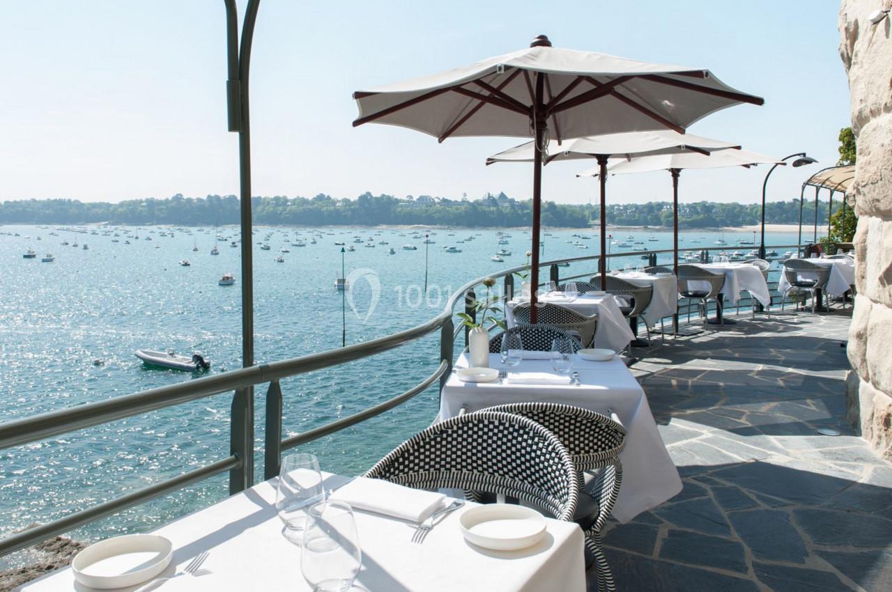 Terrasse d'un restaurant en bord de mer avec tables dressées, parasols et vue sur un paysage marin parsemé de bateaux.