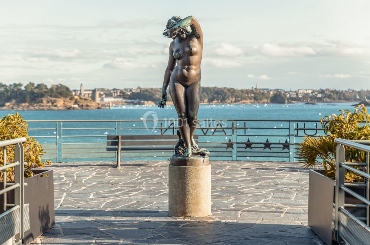 Statue en bronze représentant une femme nue, située sur une terrasse avec vue sur la mer et un paysage côtier.
