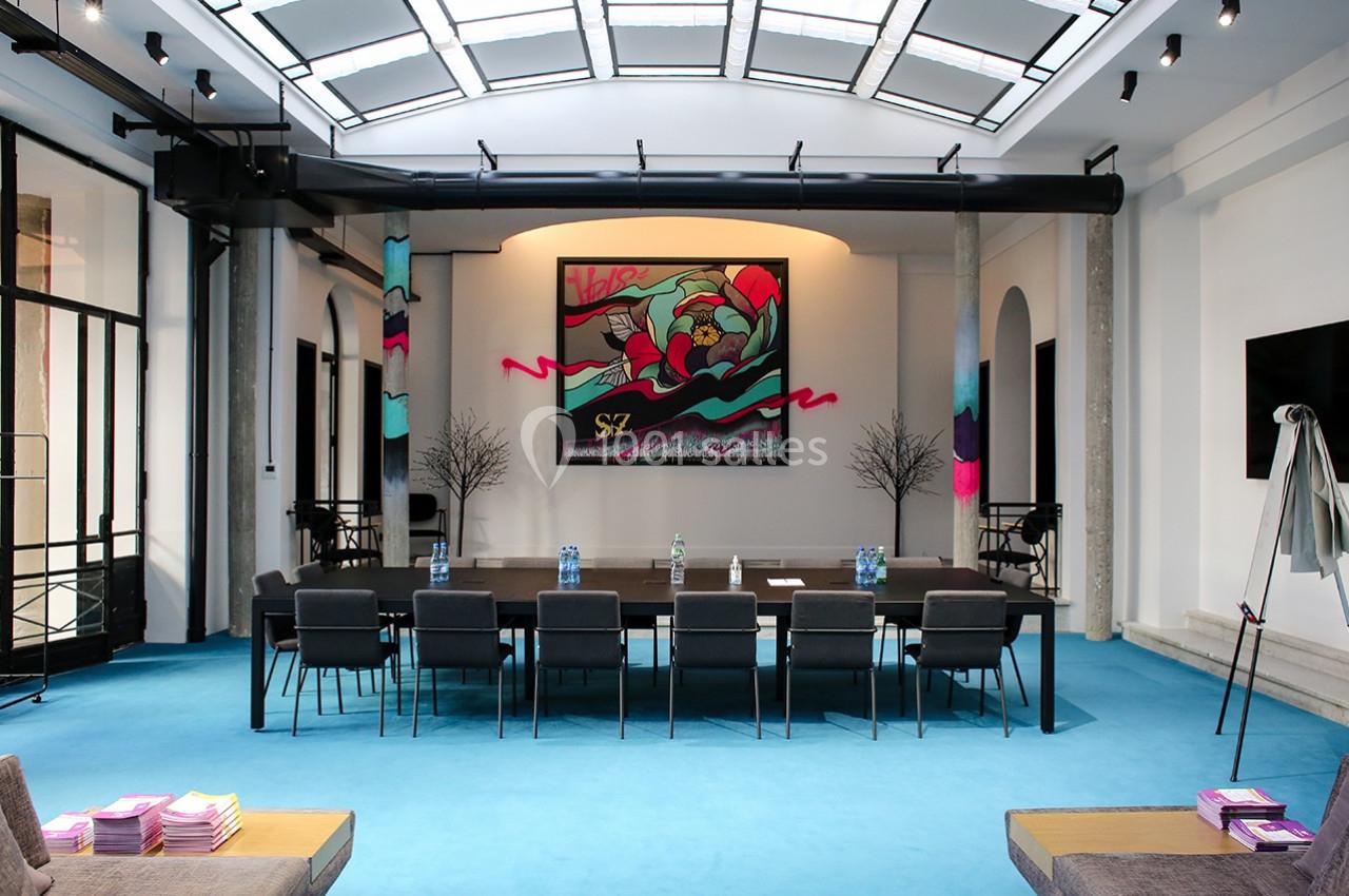 Salle de réunion moderne avec table rectangulaire, chaises noires, sol bleu et tableau coloré en arrière-plan.