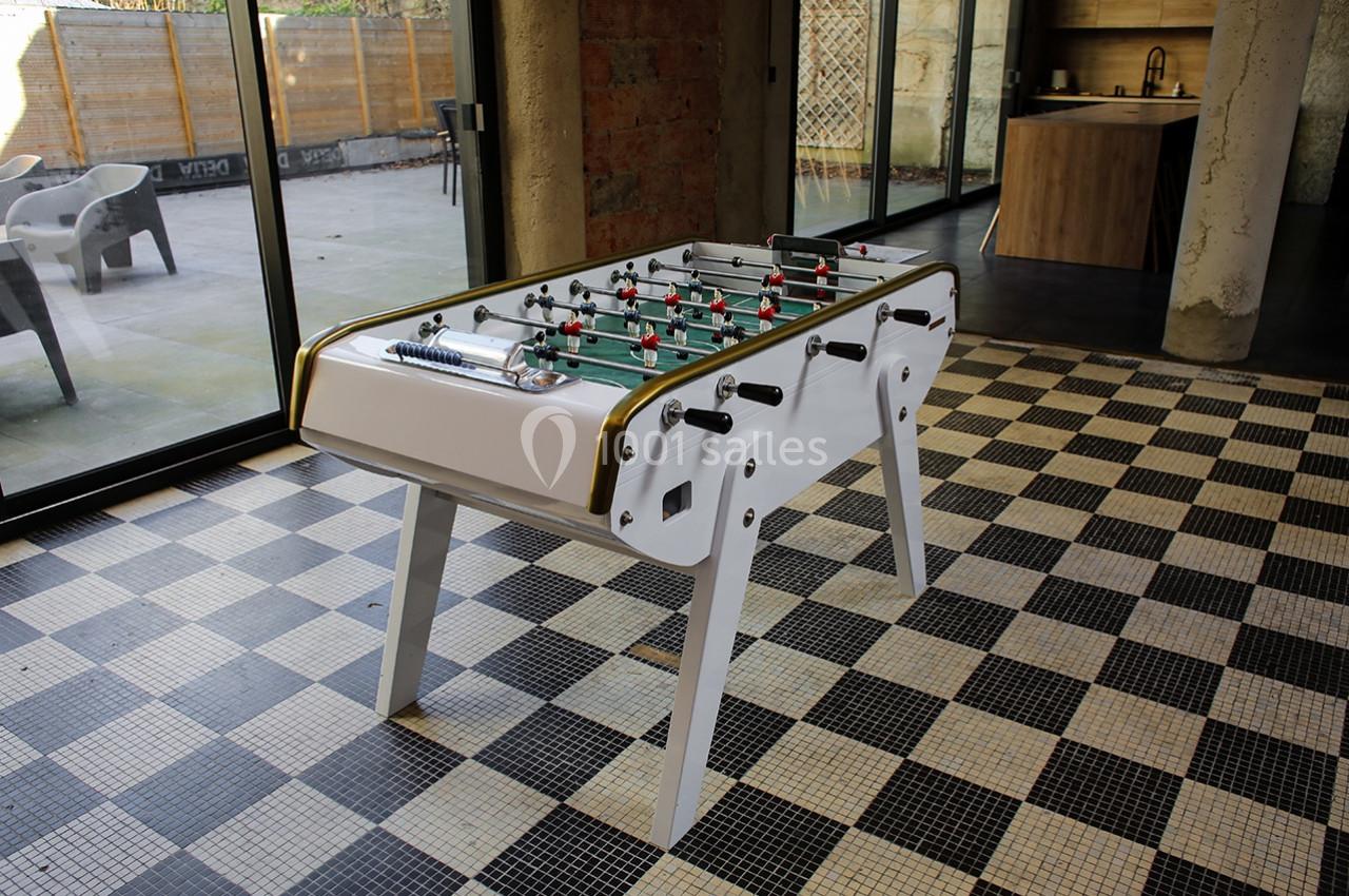 Table de baby-foot blanche sur un sol en carreaux noirs et blancs, dans une pièce lumineuse avec vue sur une terrasse.