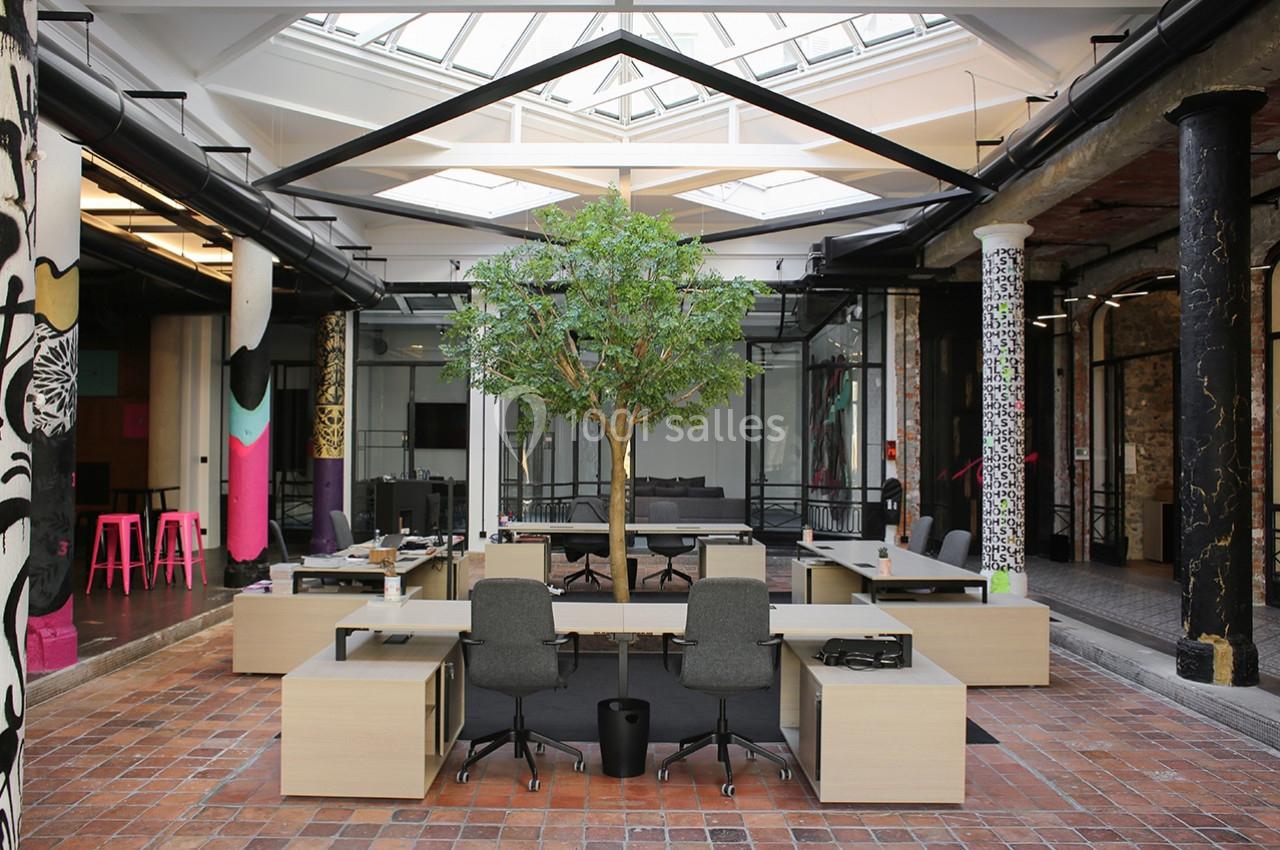 Espace de travail moderne avec bureaux partagés, un arbre central et une verrière laissant entrer la lumière naturelle.