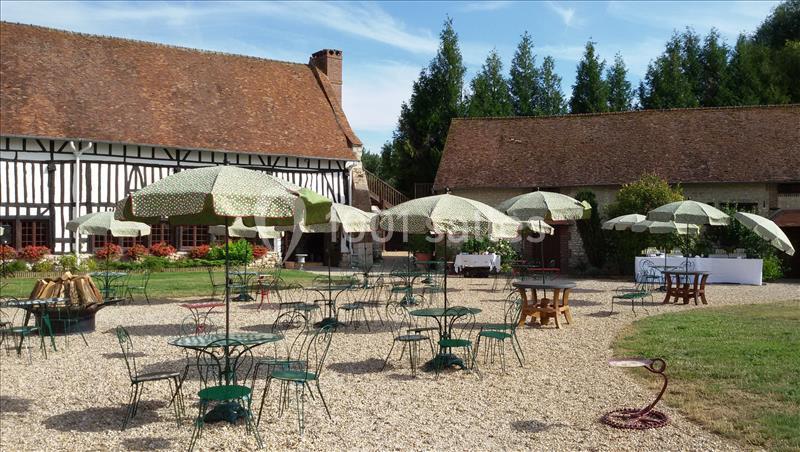 Cour extérieure avec tables et chaises en fer forgé sous des parasols, entourée de bâtiments à colombages et de verdure.