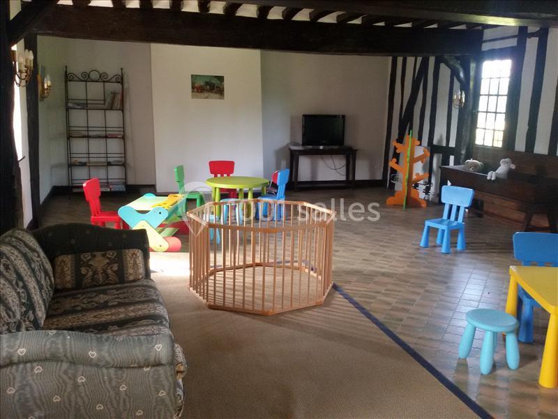 Salle de jeux pour enfants avec chaises colorées, tables, parc en bois et canapé dans un espace lumineux au sol carrelé.