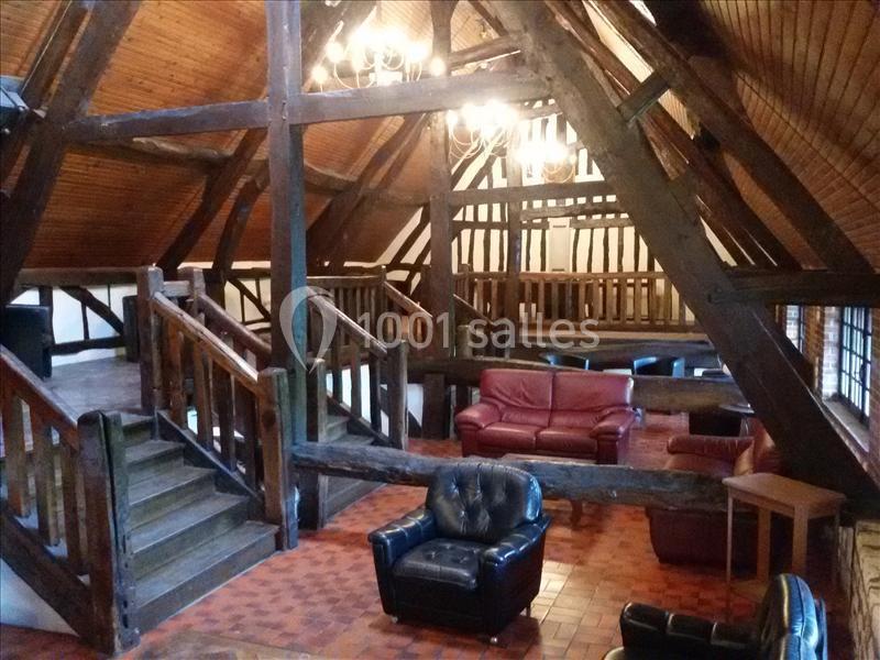 Intérieur d'une pièce avec poutres apparentes, mobilier en cuir et escalier en bois, éclairée par des lustres.