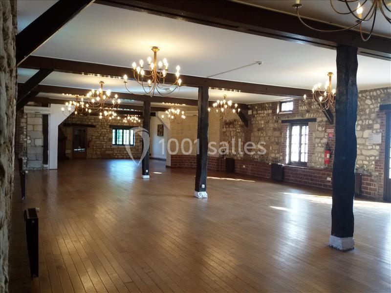 Salle spacieuse avec parquet en bois, murs en pierre apparente, poutres sombres et lustres suspendus.