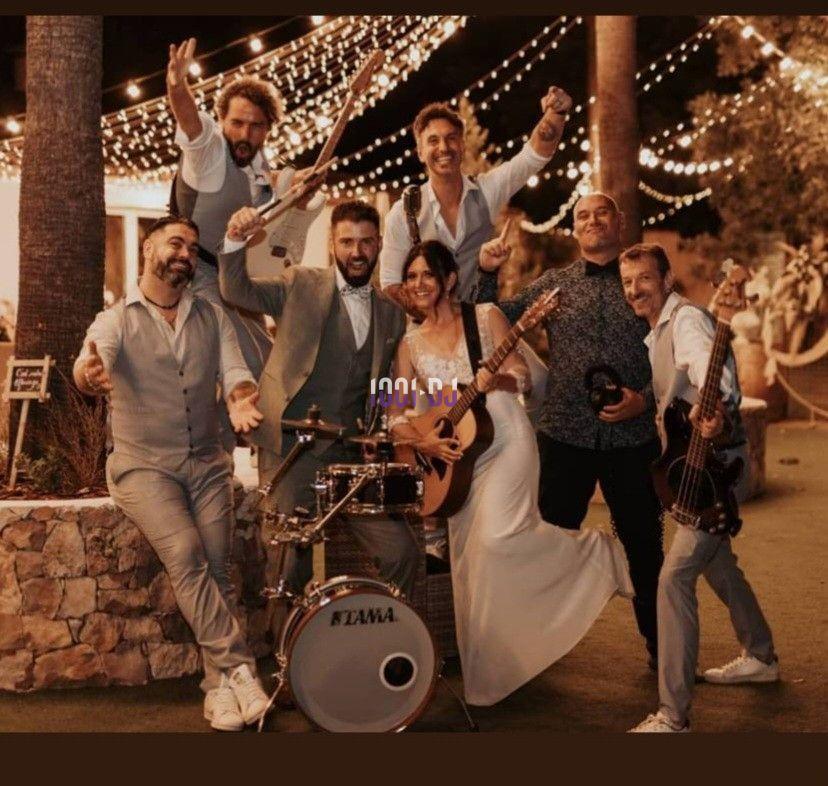 Un groupe de musiciens et une femme en robe blanche posent joyeusement avec leurs instruments sous des guirlandes lumineuses.