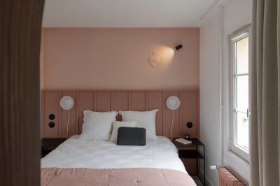 Chambre moderne avec lit double, mur rose, bureau en bois clair, chaise noire et éclairage mural.