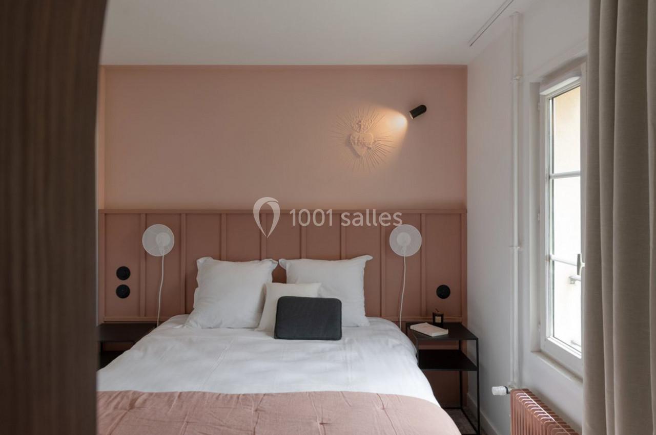 Chambre lumineuse avec lit double, tête de lit rose, lampes murales et fenêtre laissant entrer la lumière naturelle.