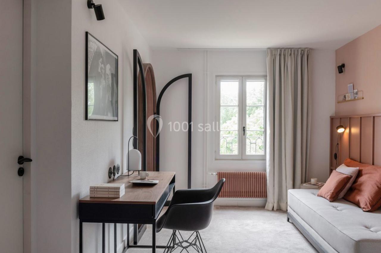 Bureau avec chaise noire, lampe et livres, dans une chambre lumineuse avec canapé, rideaux clairs et fenêtre ouverte.