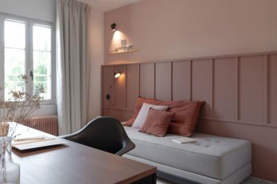 Chambre moderne avec lit double, mur rose, bureau en bois clair, chaise noire et éclairage mural.