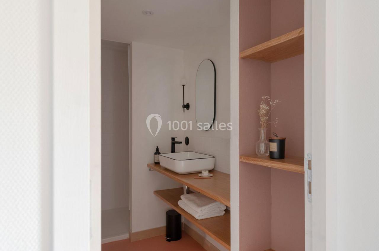 Salle de bain minimaliste avec lavabo blanc, miroir ovale, étagères en bois et mur rose pâle.