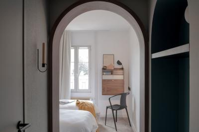 Chambre moderne avec lit double, mur rose, bureau en bois clair, chaise noire et éclairage mural.