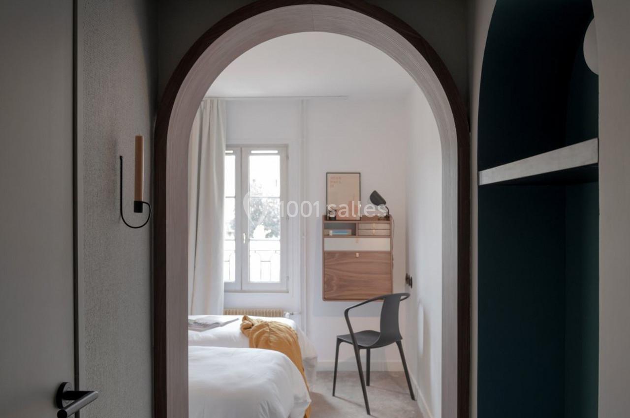 Vue d'une chambre lumineuse avec un lit, une chaise, un bureau mural en bois et une fenêtre encadrée de rideaux clairs.