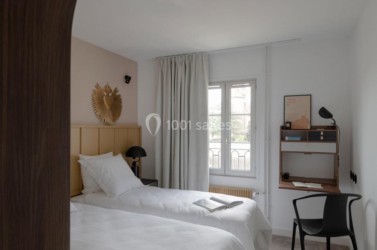 Deux lits simples avec linge blanc dans une chambre lumineuse, bureau en bois, chaise noire et fenêtre avec rideaux.