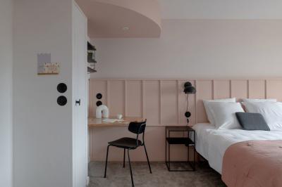 Chambre moderne avec lit double, mur rose, bureau en bois clair, chaise noire et éclairage mural.