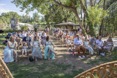 Une femme lit un discours devant un couple assis sous une arche décorée, lors d'une cérémonie en extérieur.