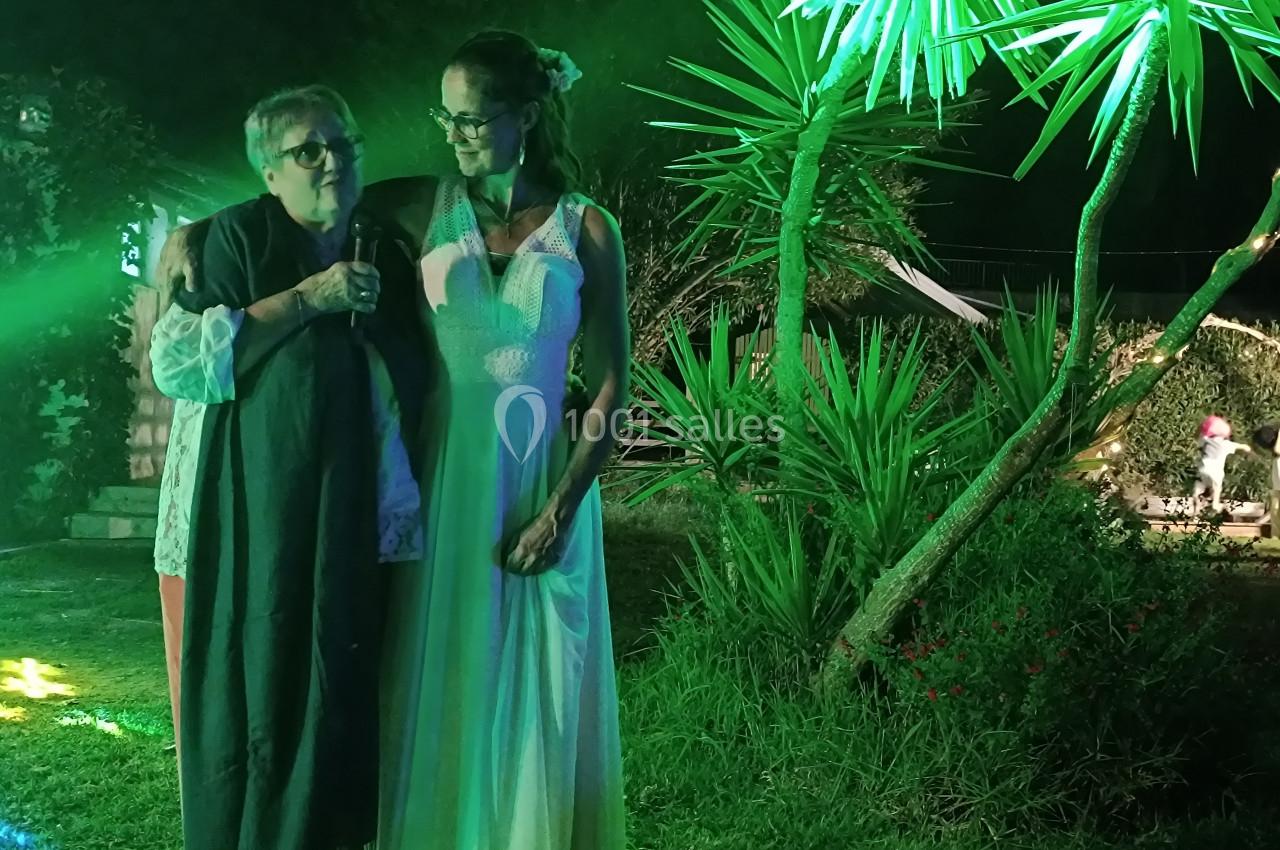 Deux femmes debout dans un jardin éclairé de lumières vertes, l'une tenant un micro, l'autre en robe blanche.