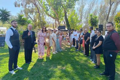 Une femme lit un discours devant un couple assis sous une arche décorée, lors d'une cérémonie en extérieur.