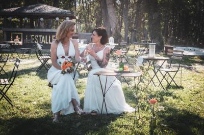 Une femme lit un discours devant un couple assis sous une arche décorée, lors d'une cérémonie en extérieur.