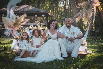 Une femme lit un discours devant un couple assis sous une arche décorée, lors d'une cérémonie en extérieur.