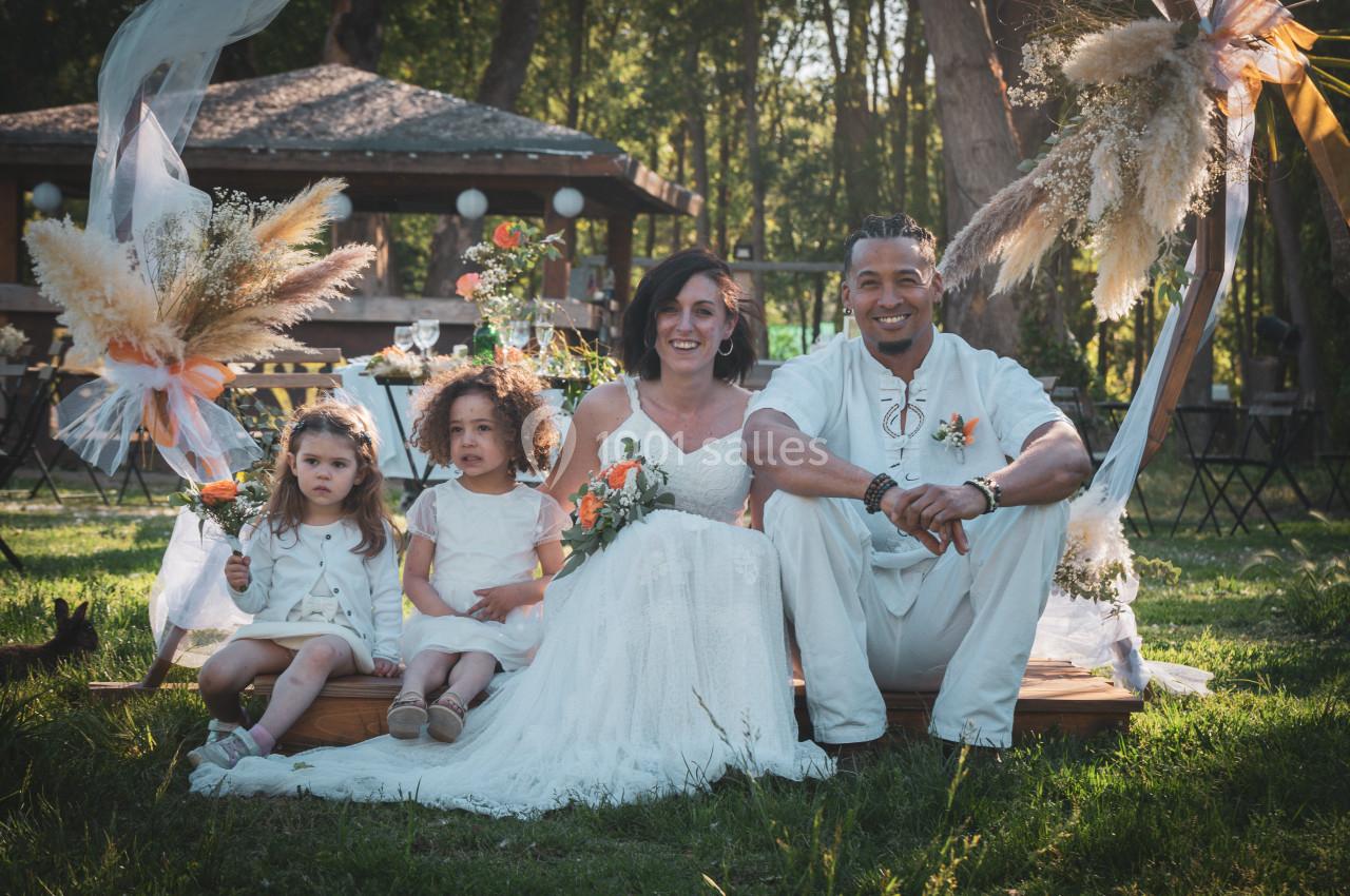 Un couple en tenue de mariage assis sur une pelouse avec deux enfants, entourés de décorations florales en extérieur.