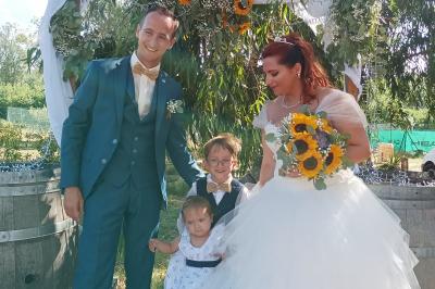 Une femme lit un discours devant un couple assis sous une arche décorée, lors d'une cérémonie en extérieur.