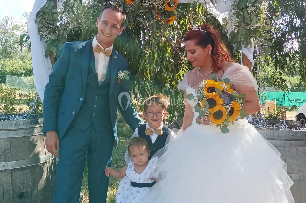Un couple en tenue de mariage pose avec deux enfants devant une arche décorée de tournesols en extérieur.