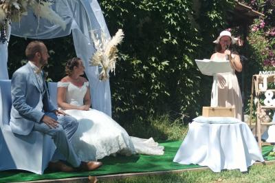 Une femme lit un discours devant un couple assis sous une arche décorée, lors d'une cérémonie en extérieur.