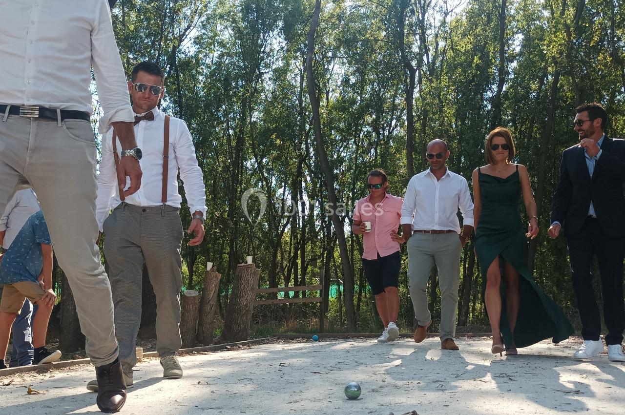 Des personnes élégamment vêtues jouent à la pétanque sur un terrain ombragé par des arbres.