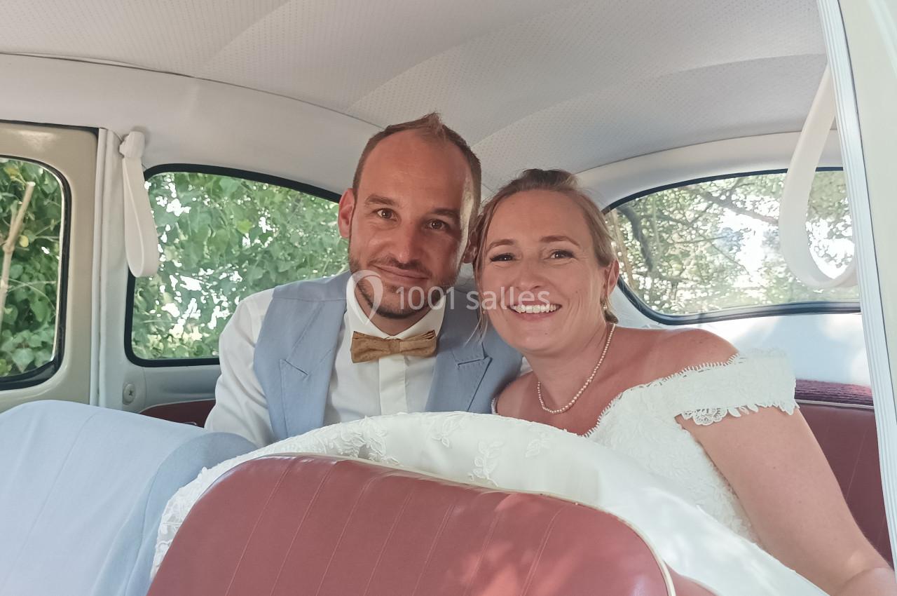 Un couple souriant en tenue de mariage assis à l'arrière d'une voiture ancienne avec un intérieur rétro.