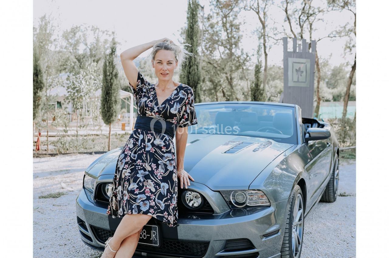 Femme en robe fleurie posant devant une voiture décapotable grise, dans un environnement extérieur arboré.