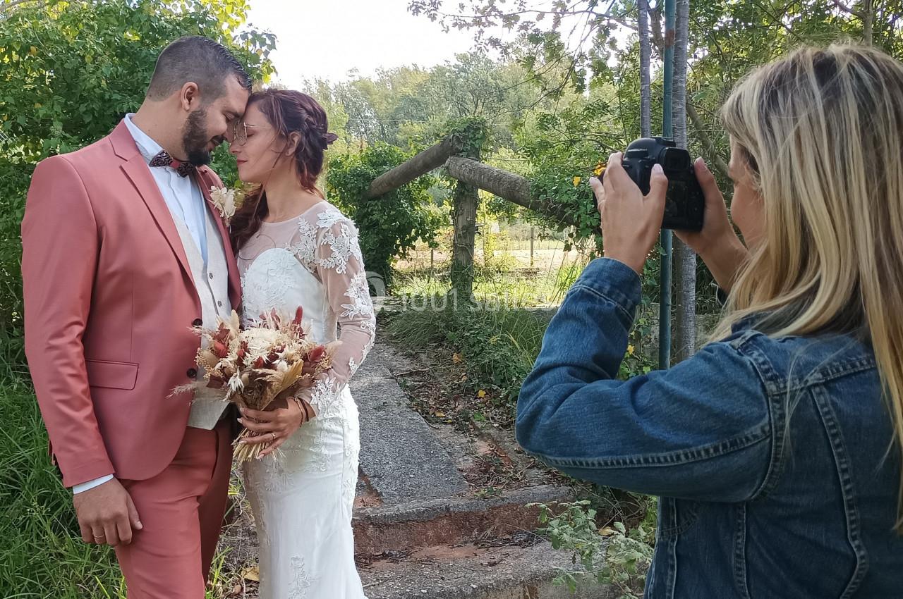 Une photographe capture un couple de mariés posant dans un jardin, la mariée tenant un bouquet champêtre.