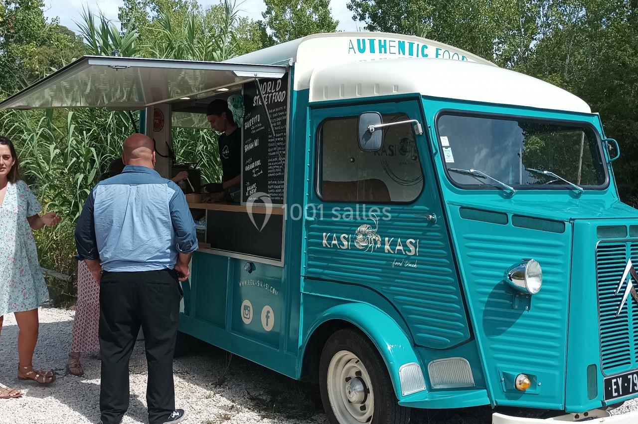 Un food truck bleu vintage avec une fenêtre de service, des clients commandant et une femme discutant à côté.