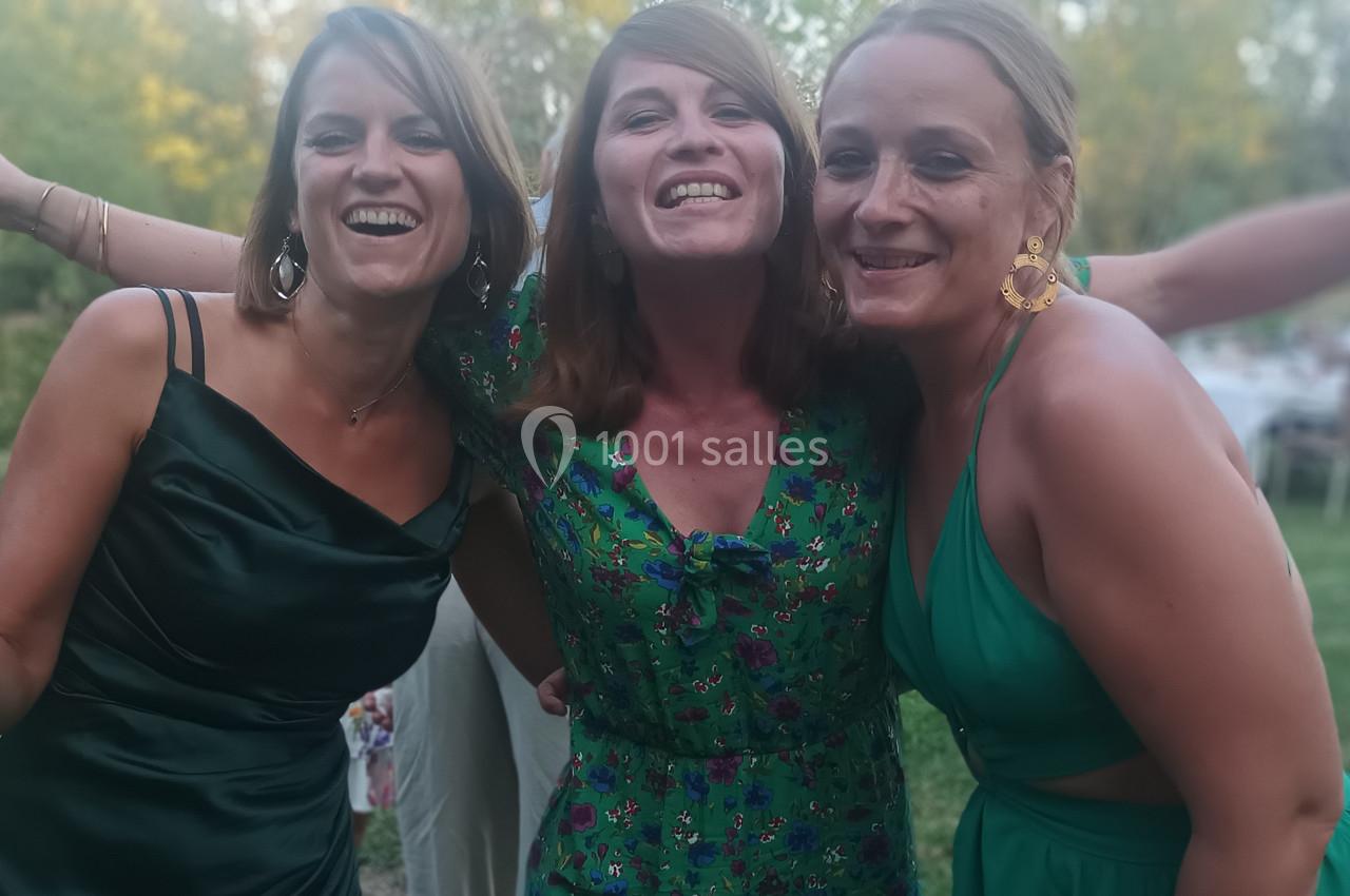 Trois femmes souriantes en tenue de soirée posent ensemble dans un jardin lors d'un événement en plein air.