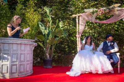 Une femme lit un discours devant un couple assis sous une arche décorée, lors d'une cérémonie en extérieur.