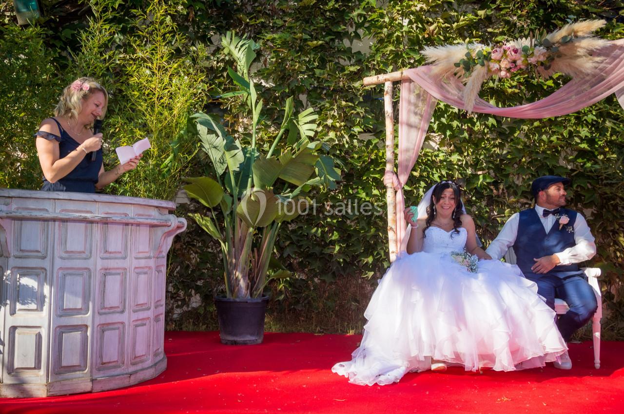 Une femme lit un discours devant un couple assis sous une arche décorée, lors d'une cérémonie en extérieur.