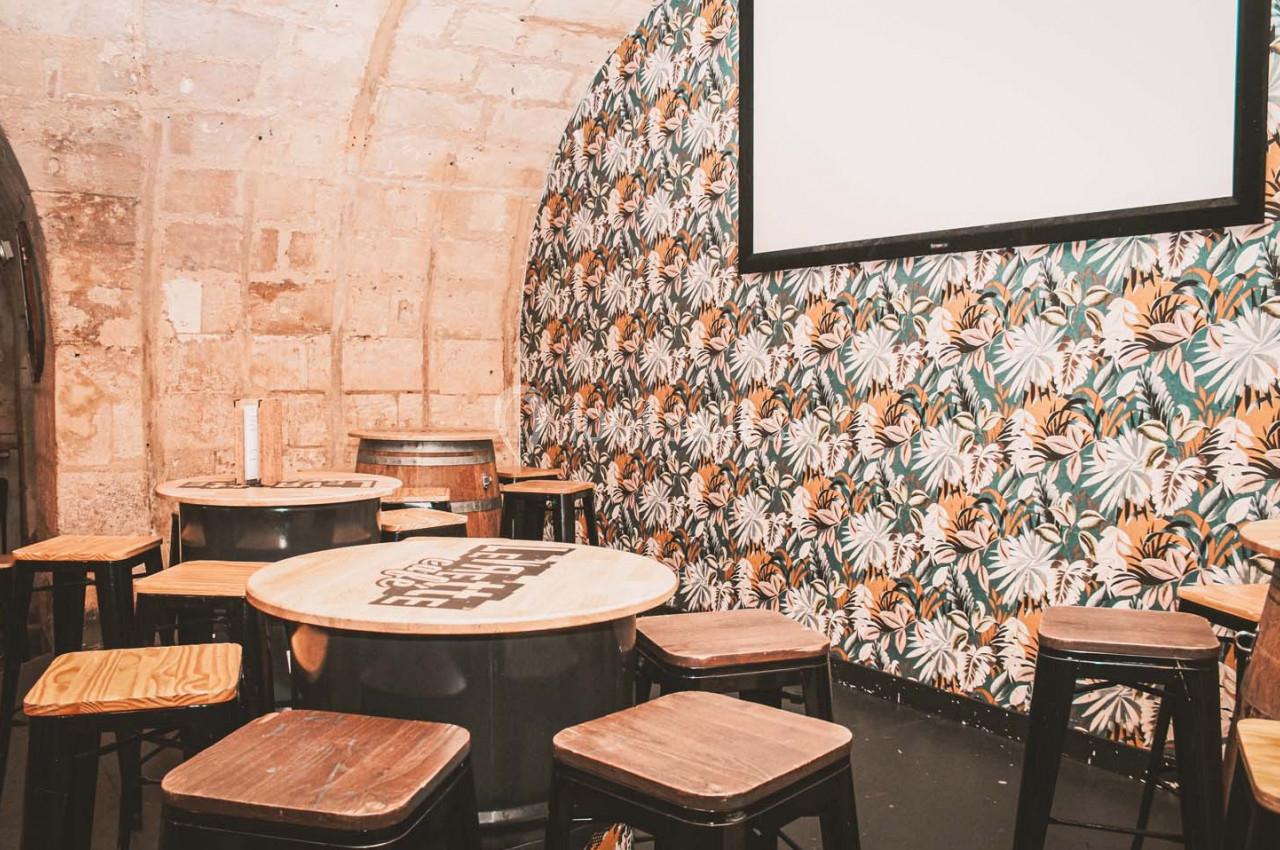 Salle avec murs en pierre et papier peint floral, équipée de tables rondes en bois, tabourets et un écran mural.