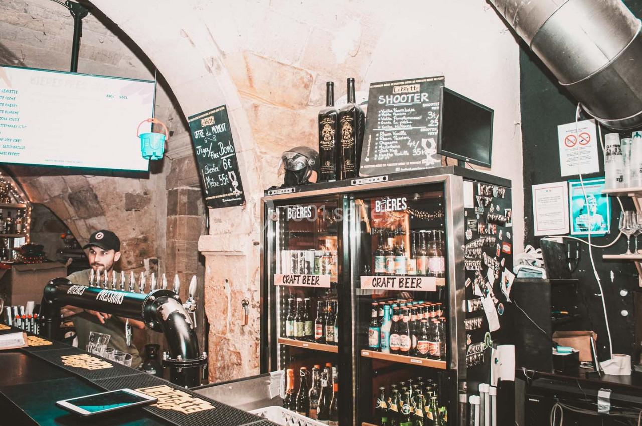 Bar avec comptoir, tireuses à bière et réfrigérateurs remplis de bouteilles de bières artisanales dans un décor en pierre.
