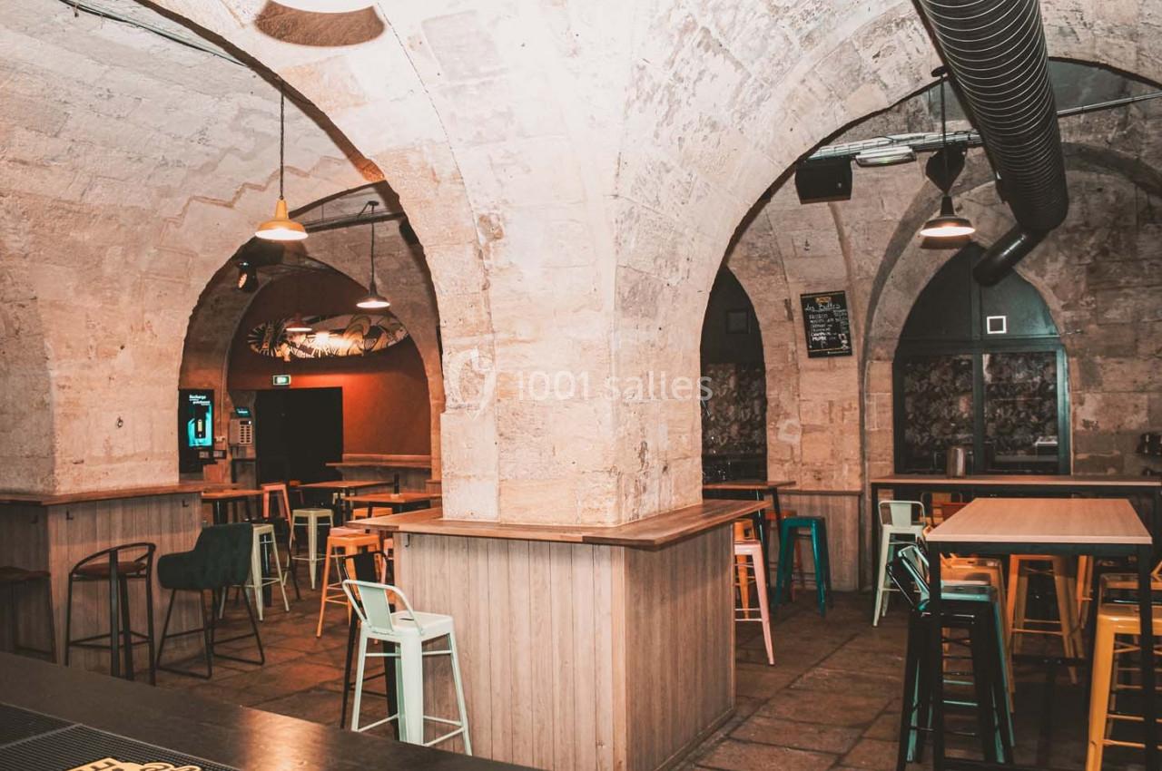 Salle voûtée en pierre avec tables hautes, chaises colorées et éclairage tamisé.