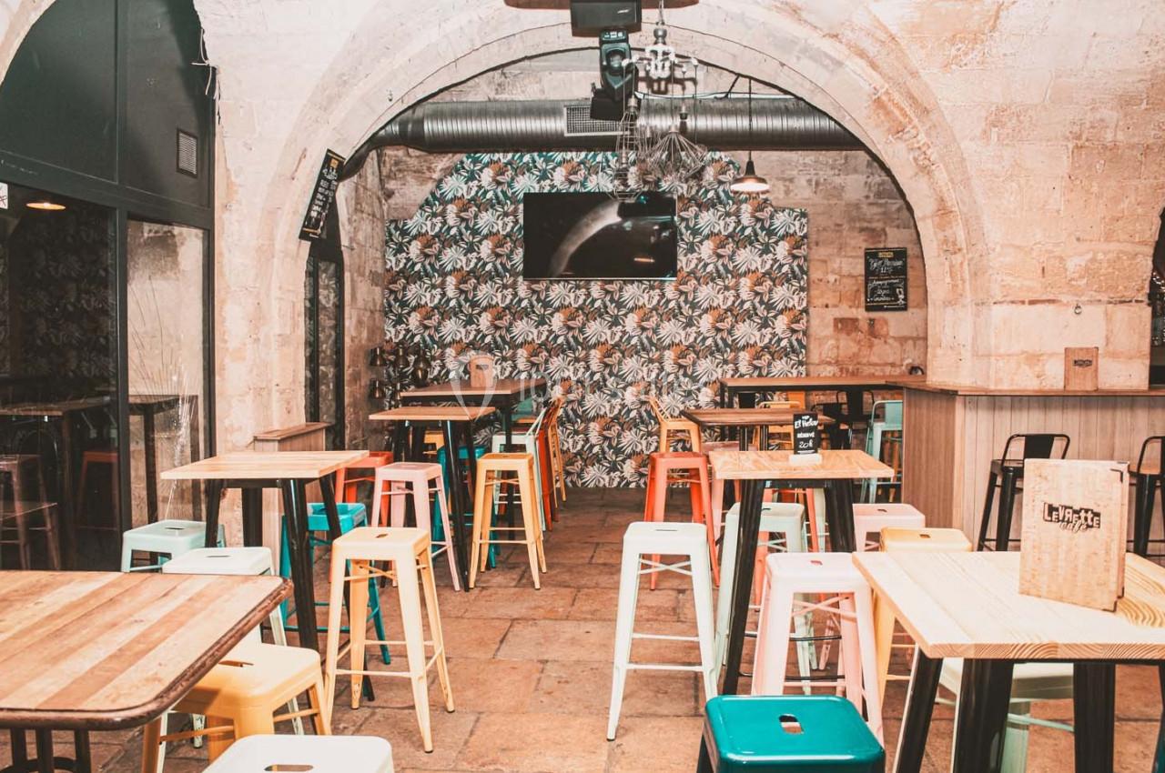Salle de bar avec tables en bois, tabourets colorés et décor mural à motifs tropicaux sous une voûte en pierre.