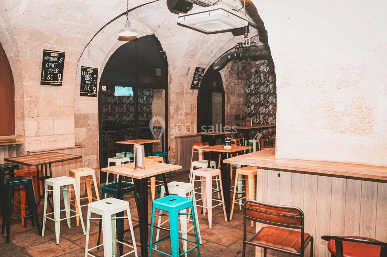 Salle de bar avec tables hautes, tabourets colorés et murs en pierre voûtés.